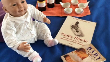 Eine Puppe sitzt auf einer blauen Unterlage, umgeben von Massageölen, Schalen und zwei Büchern über Babymassage.
