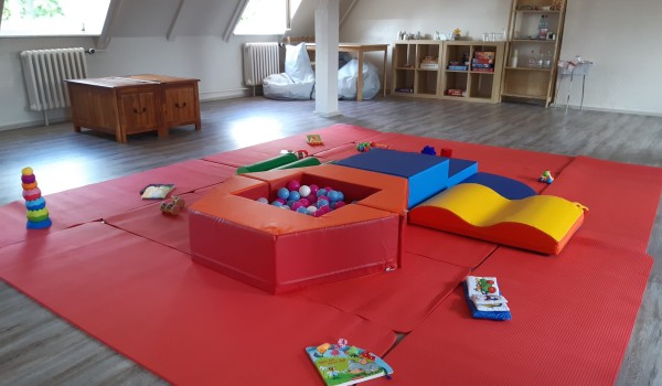 Bunte Spielmatte mit verschiedenen Spielzeugen, darunter ein Ballbecken, Kissen und Bauklötze in einem hellen Raum.