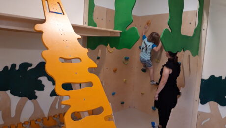 Ein Kind klettert an einer bunten Kletterwand, während eine Erwachsene es beobachtet und unterstützt.