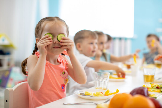 Kinder beim Essen. Mädchen im Vordergrund hält sich Gurkenscheiben vor die Augen