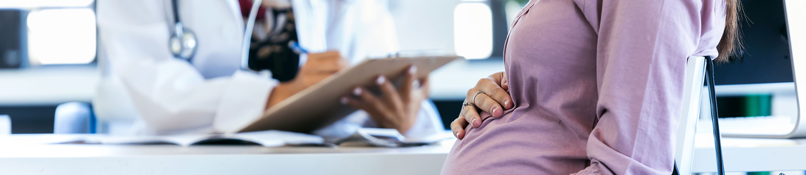 Titelbild - Schwangere Frau sitzt am Schreibtisch eines Arztes und hält die rechte Hand auf ihren Bauch