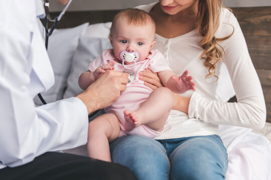 Arzt hört Baby ab, welches auf dem Schoß der Mutter sitzt