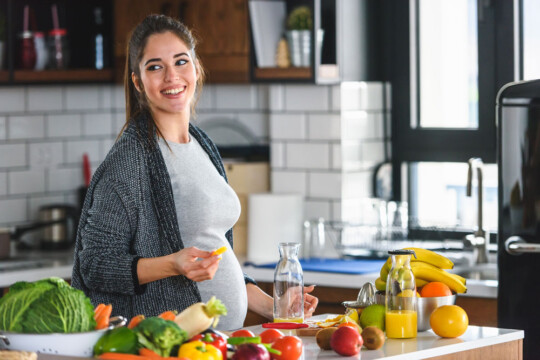 Schwangere Frau steht lächelnd in der Küche und macht sich Frühstück