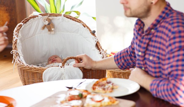Ein schlafendes Baby in einem Weidenkorb, ein Vater isst Pizza am Tisch.