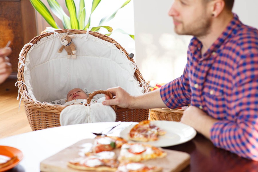 Ein schlafendes Baby in einem Weidenkorb, ein Vater isst Pizza am Tisch.