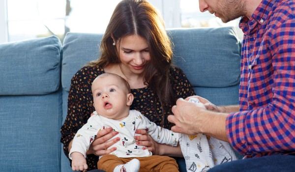 Mutter sitzt mit Baby auf der Couch und Vater hockt davor und hilft beim Anziehen des Babys