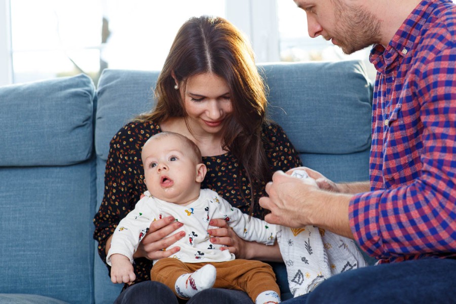 Mutter sitzt mit Baby auf der Couch und Vater hockt davor und hilft beim Anziehen des Babys