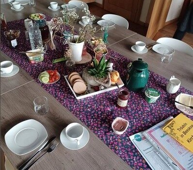 Tisch mit Frühstücksbuffet: Gebäck, Obst, Getränke, Geschirr und Blumenschmuck auf blumigem Tischläufer.