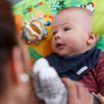 Ein Baby liegt auf einer bunten Spielmatte und blickt neugierig auf einen Erwachsenen, der ihm ein Spielzeug zeigt.